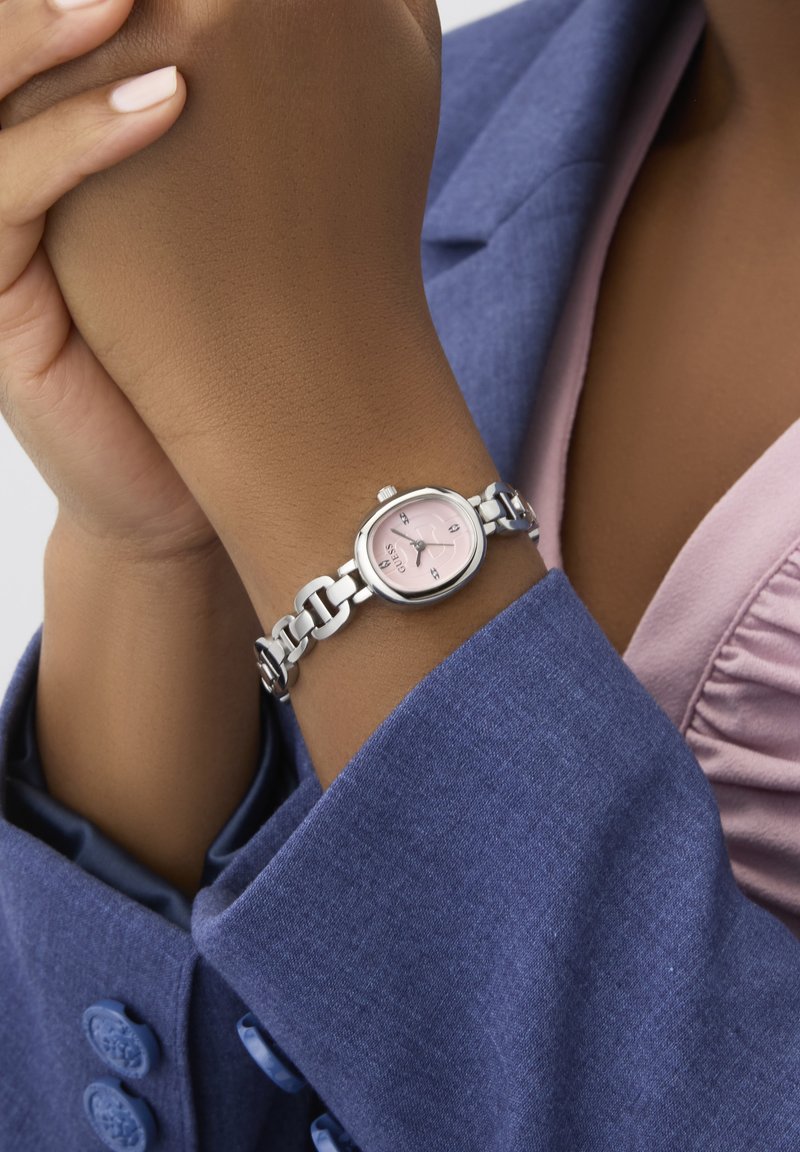 Zilveren horloge met een roze wijzerplaat, ovale wijzerplaat en een armband van schakels. Draagt het om een pols met een donkere huidskleur, tegen een blauwe stoffen achtergrond.