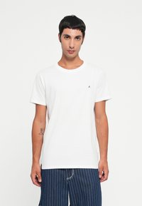 Tricou alb din bumbac, cu decolteu rotund și mâneci scurte. Are un mic logo negru pe pieptul stâng și o textură netedă.