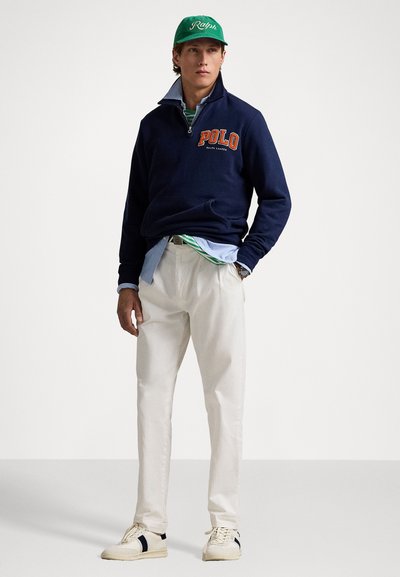 Sudadera con cremallera en color azul marino con "POLO" en naranja, camisa azul claro, pantalones blancos y gorra verde. El calzado es de color crema con acentos en azul marino.