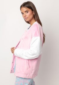 Chaqueta bomber de felpa rosa con mangas blancas, botones a presión y bordado en la letra. Textura suave, colores contrastantes y diseño oversize.