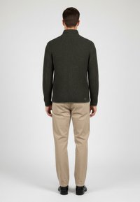 Maglione a coste di un verde scuro con collo alto, stile aderente, indossato con pantaloni beige e scarpe nere, sfondo neutro, vista posteriore.