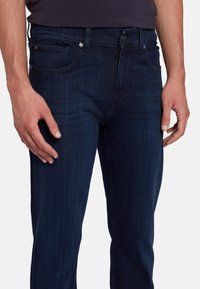 Jeans in denim blu scuro presentano un design classico con gamba dritta, stile a cinque tasche e dettagli in metallo nella fascia in vita.