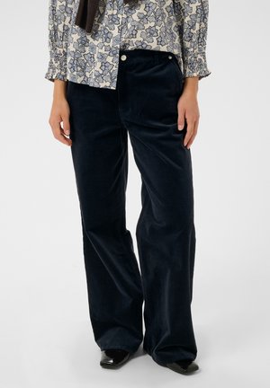 Pessoa vestindo calças largas de veludo preto combinadas com uma blusa leve floral de manga comprida e sapatos pretos pontiagudos.