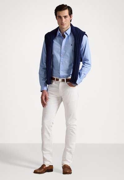 Jūras zilas adītas vestes virsma ar gaiši zilu pogājamā krekla, baltu biksu un brūnu apavu kombināciju. Apģērbs izceļ klasisku dizainu ar vienkāršām akcentiem.