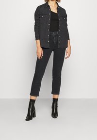 Mujer con jeans cortos negros, botines negros, una blusa negra de un solo hombro y una chaqueta de mezclilla negra desabrochada.