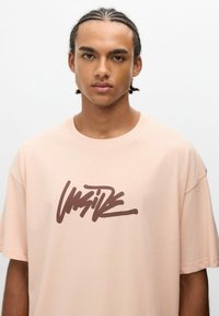 Camiseta de gran tamaño en color rosa claro, hecha de algodón, con un destacado logotipo en estilo graffiti marrón en el centro. Mangas cortas y escote redondo.