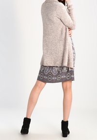 Jasnobrązowy sweter z dzianiny z kołnierzem szalowym, zestawiony z szaro wzorzystą sukienką. Czarne botki na kostkę dopełniają cały strój.