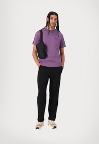 Camiseta de algodón morada con logotipo, pantalones jogger negros y zapatillas beige. Accesorio: pequeña mochila cruzada negra con cremallera.