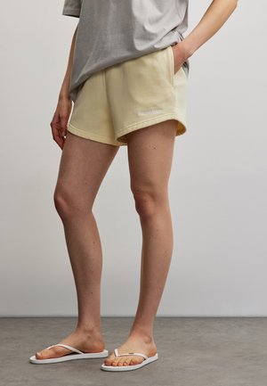 Femme portant un short beige et un t-shirt gris, la main dans la poche, debout pieds nus en tongs blanches sur un sol gris devant un mur uni.