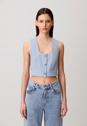 Donna con capelli lisci e scuri indossa un top corto senza maniche azzurro chiaro con bottoni e jeans a vita alta a lavaggio chiaro, in piedi davanti a uno sfondo semplice.