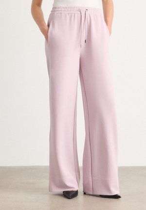 Pantalon de jogging lilas à jambes larges avec taille élastique et cordon, porté avec des chaussures noires à bout pointu, mains dans les poches.