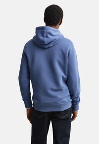 Mand iført en blå hættetrøje og mørke jeans, stående med ryggen mod en ensfarvet lys baggrund.