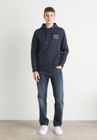 Marineblå hettegenser med frontlomme og snorer, med en liten hvit logo. Kombinert med mørkeblå jeans og hvite sneakers.