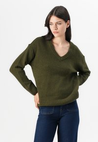 Oliven grønn v-hals genser med ribbestrikkede mansjetter og kant, laget av et mykt strukturert materiale, kombinert med mørkeblå jeans.