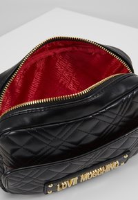 Svart quiltad handväska med gyllene detaljer, med en klar röd foderinteriör som visar ett upprepat logomönster. Dragkedjestängning synlig.