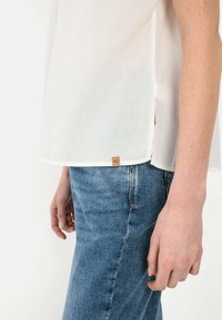 Weißes, leichtes Hemd mit kurzen Ärmeln, abgerundetem Saum und seitlichem Schlitz; hat ein kleines braunes Stoffetikett an der unteren Seite, kombiniert mit blauen Jeans.