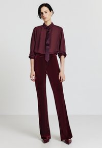 Blusa di seta bordeaux con un nodo e maniche lunghe, abbinata a pantaloni in velluto a vita alta coordinati, caratterizzati da una superficie liscia e gambe larghe.