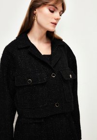 Schwarze, cropped Jacke aus strukturiertem Stoff mit metallischen Akzenten. Verfügt über zwei Fronttaschen und einen Reverskragen. Glatter, strukturierter Schnitt.