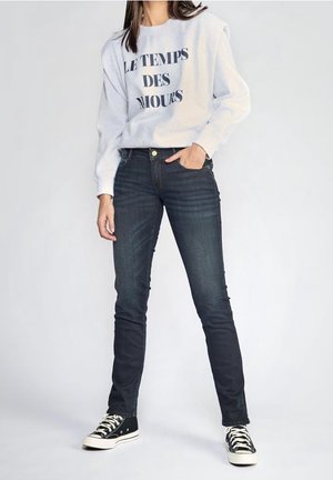 Osoba ubrana w białą bluzę z nadrukiem "LE TEMPS DES AMOURS", ciemne dopasowane jeansy i czarno-białe trampki, stojąca na tle jednolitego tła.