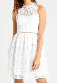 Vitt spetsklänning med hög hals, ärmlös design, texturerade blommönster och en utsvängd kjol med en subtil fålldetalj.