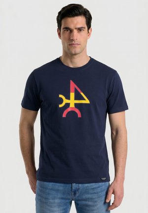 Hombre con camiseta azul marino con diseño geométrico abstracto en amarillo y rojo, combinado con vaqueros azules, de pie frente a un fondo liso.