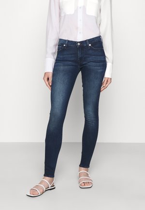 Slim fit donkerblauwe spijkerbroek gedragen door een persoon met een wit overhemd met knopen en witte sandalen met bandjes op een effen achtergrond.
