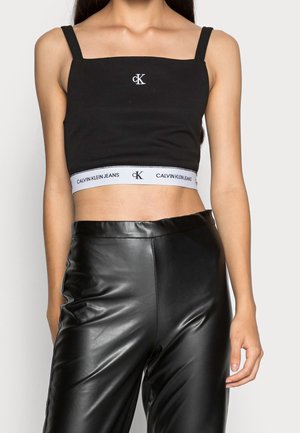 Femme portant un crop top Calvin Klein noir avec logo et un pantalon en cuir noir, montrée des épaules aux hanches sur un fond uni.