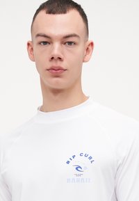 Hvit langermet rash guard med raglanermer, med en blå bølgedesign og teksten "RIP CURL HAWAII" i variert typografi.