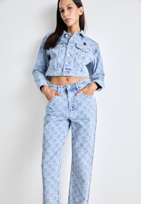 Chaqueta de mezclilla azul claro con un corte cuadrado, que cuenta con bolsillos frontales y cierre de botones. Pantalones azul claro a juego con un patrón de logotipo repetido.
