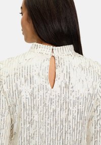 Weißes Kleid mit hohem Ausschnitt, strukturiertem Stoff, der vertikale metallische Fäden aufweist, und einem Schlüsselelement am Rücken, das mit Knöpfen gesichert ist.