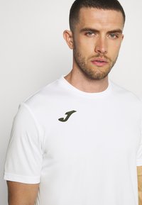 Bílý sportovní shirt vyrobený z hladké tkaniny, s kulatým výstřihem a tmavě zeleným logem na levé horní straně.