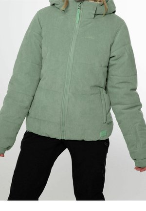 Kobieta ubrana w jasnobedrową pikowaną kurtkę ze sztruksu z kapturem i czarne spodnie, stojąca z lekko uniesionymi ramionami.