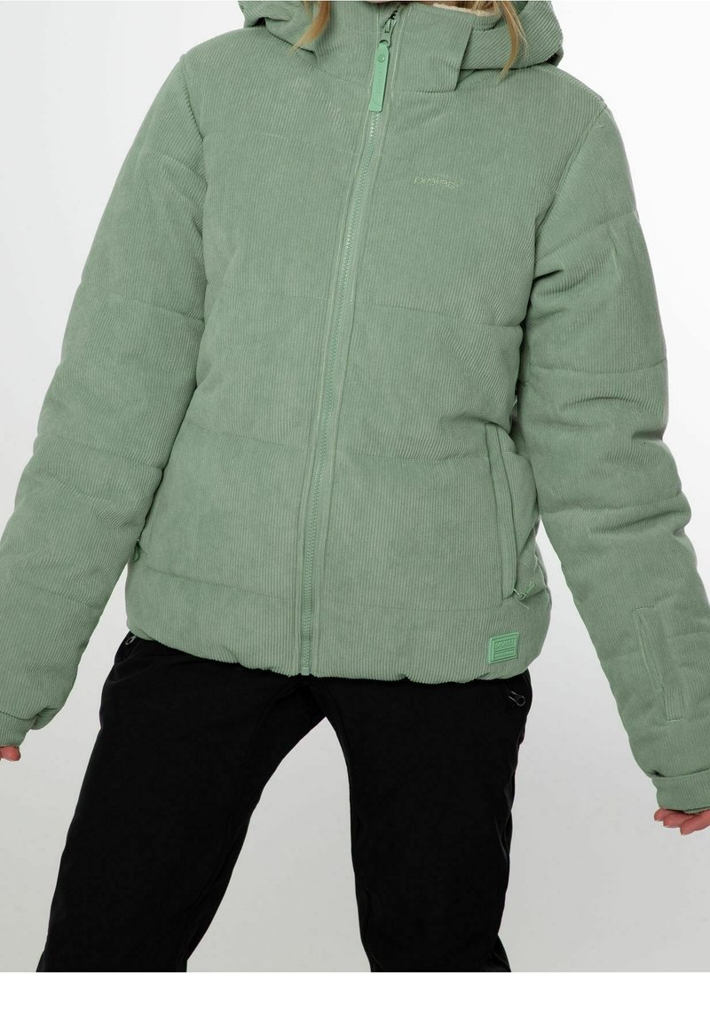 Femme portant une doudoune en velours côtelé vert clair avec une capuche et un pantalon noir, debout, les bras légèrement étendus.