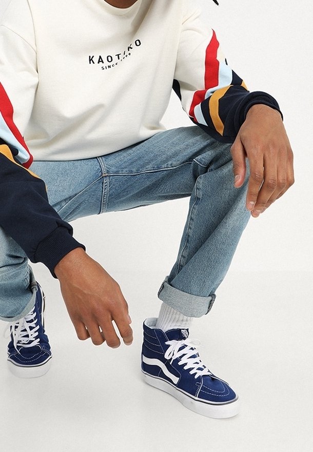 Homme portant des baskets montantes bleues, un jean bleu clair retroussé et un sweat-shirt blanc avec des bandes rouges, bleues et orange sur les manches.