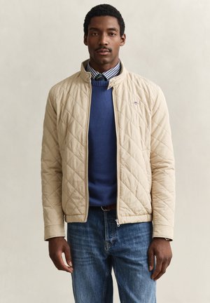 Man draagt een beige gewatteerd jack over een blauw vest, gestreept overhemd en stropdas, gecombineerd met een blauwe spijkerbroek, staande tegen een effen achtergrond.