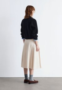 Schwarzer Strickpullover, hellblaues Hemd mit Kragen darunter, cremefarbener, plissierter Midirock mit ausgefranstem Saum, graue Socken und braune Mokassins.