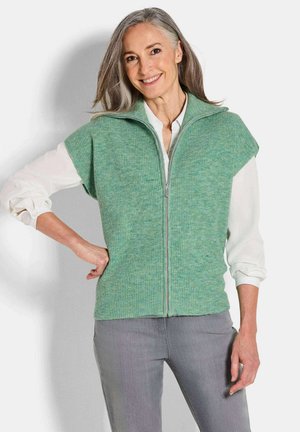 Gilet en tricot vert avec un col montant et une fermeture éclair, doté de manches courtes et d'un motif texturé. Porté sur une chemise blanche.