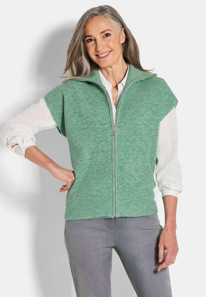 Grüne gestrickte Weste mit hohem Kragen und Reißverschluss, mit kurzen Ärmeln und einem strukturierten Muster. Über einem weißen Hemd getragen.