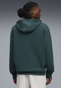 Felpa con cappuccio di un verde scuro, realizzata in materiale morbido, caratterizzata da un cappuccio ampio e polsini a coste, indossata sopra pantaloni della tuta grigi chiari.