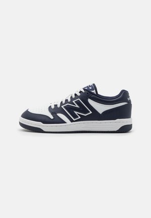 Low-top New Balance sneaker i navy blå og hvid med hvide snørebånd, perforeret tå og tyk sål på ensartet lyst baggrund.