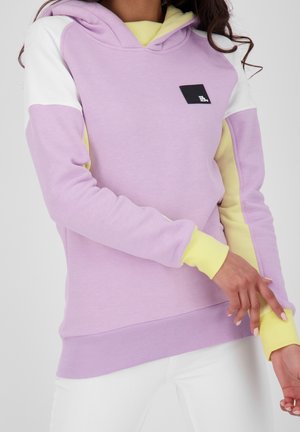 Femme portant un sweat à capuche violet pastel avec des poignets jaunes et une doublure de capuche, des panneaux blancs aux épaules et un petit patch logo noir sur la poitrine.