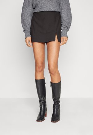 Jupe noire mini avec une fente sur le devant, associée à des bottes en cuir noir montantes avec un bout carré et un talon carré, portée avec un pull gris.