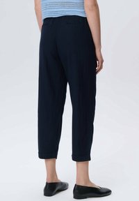 Personne portant un pantalon court navy, un haut en tricot bleu clair et des ballerines noires, se tenant devant un fond clair uni.