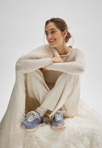 Zapatillas azules con acentos rosas, cordones blancos y suelas texturizadas. El modelo lleva un atuendo beige y una manta cremosa grande y esponjosa.
