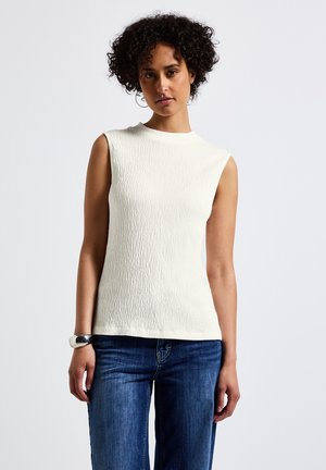 Femme aux cheveux bouclés portant un top blanc texturé sans manches, un jean bleu et un bracelet en argent sur un fond blanc uni.