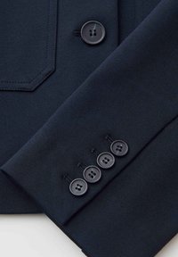 Manica di blazer blu scuro con quattro bottoni neri e parte della tasca anteriore con un singolo bottone nero su tessuto liscio.