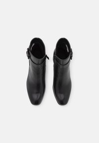 Botas de cuero negro hasta el tobillo con textura suave, punta afilada y cierre lateral con cremallera. Cuenta con una hebilla decorativa en la correa del tobillo.