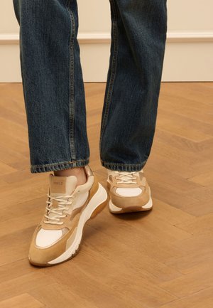 Beige and white sneakers with a textured surface, chunky sole, and laces, paired with dark blue denim jeans on a wooden floor.