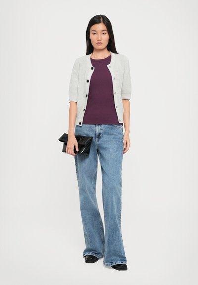Lys grå ribbestrikket cardigan med svarte knapper, parret med en lilla topp og lyseblå svakt utsvingte jeans. Holder en svart clutch-veske.