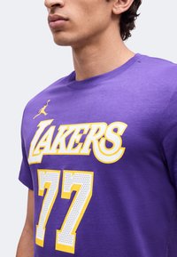 Νεαρός άνδρας φοράει μια μωβ μπλούζα Los Angeles Lakers με τον αριθμό 77 σε άσπρο και κίτρινο μπροστά.